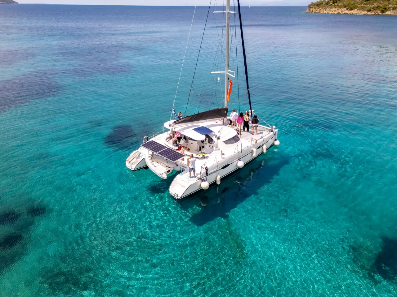 High-angle aerial drone view looking down at a modern white cruising catamaran safely anchored in a pristine, shallow turquoise bay in Halkidiki, Greece. About ten happy travelers are spread across the expansive decks and trampolines. The water is exceptionally clear, and the coastline is lush with green pine trees. A Greek flag is visible on the mast